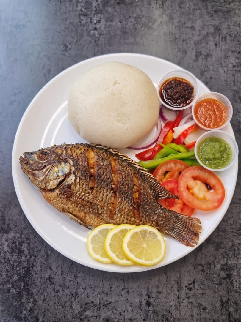 Fried whole fish with fufu, sliced vegetables, lemon, and three dipping sauces on a white plate.