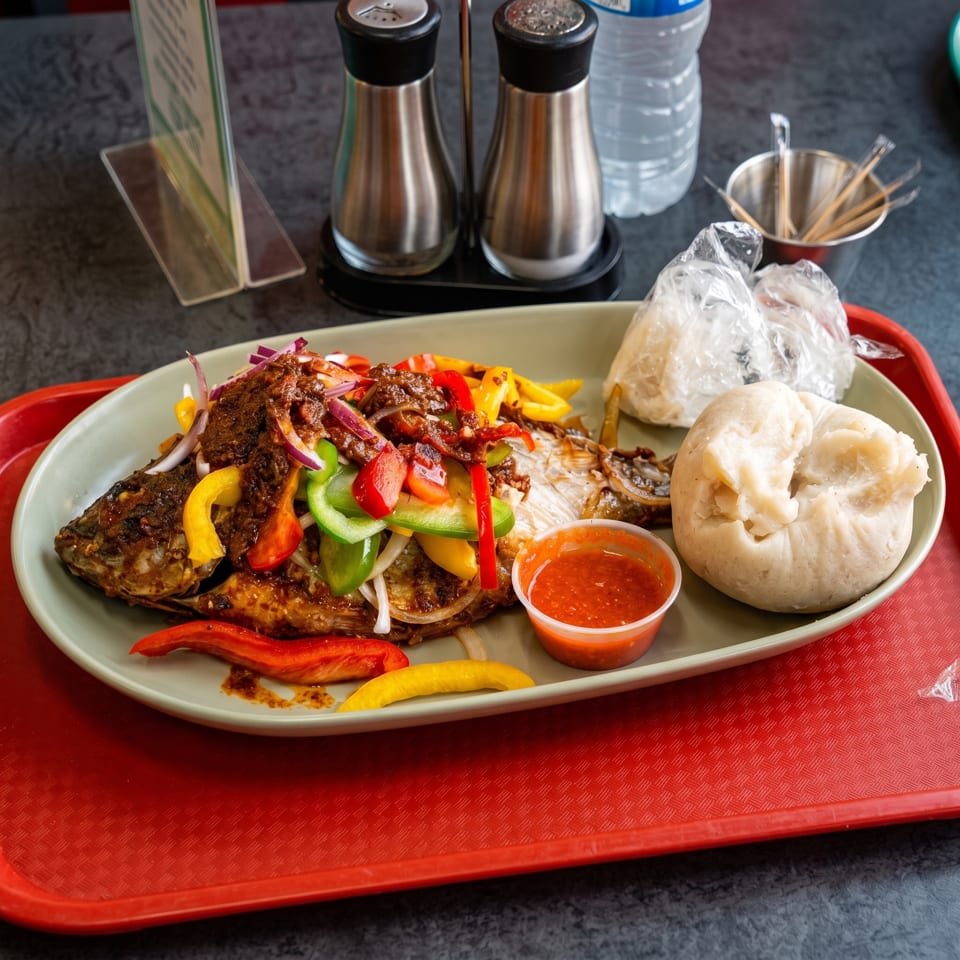 Grilled whole fish topped with colorful bell peppers, onions, and sauce, served with fufu and hot sauce.