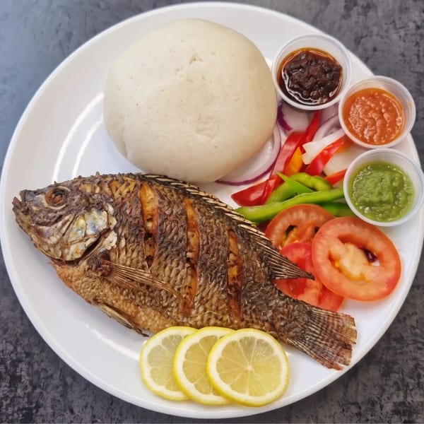 Whole fried fish with fufu, lemon slices, fresh vegetables, and three dipping sauces on a white plate.